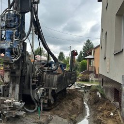 OZE-ELECTRA - Wiertnica do odwiertów geotermalnych przy budynku mieszkalnym. W tle widoczny wykop pod instalację. Pochmurne niebo.