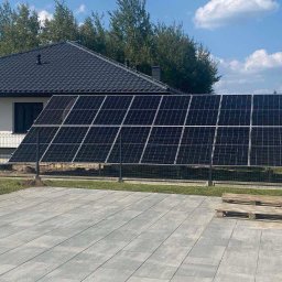 OZE-ELECTRA - Panele fotowoltaiczne zamontowane na gruncie, widoczne z boku domu z ciemnym dachem. Na pierwszym planie szara kostka brukowa i palety. Słoneczny dzień.