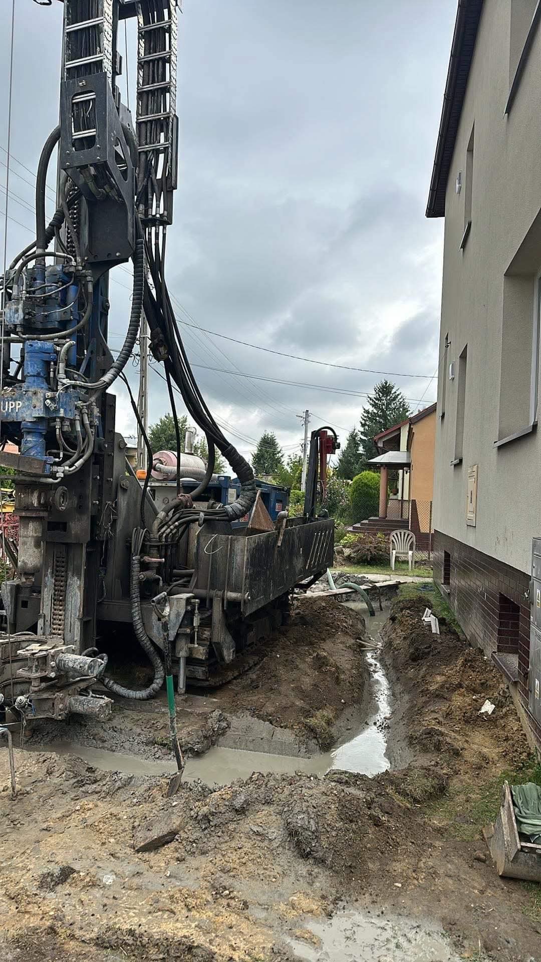 Wiertnica do odwiertów geotermalnych przy budynku mieszkalnym. W tle widoczny wykop pod instalację. Pochmurne niebo.