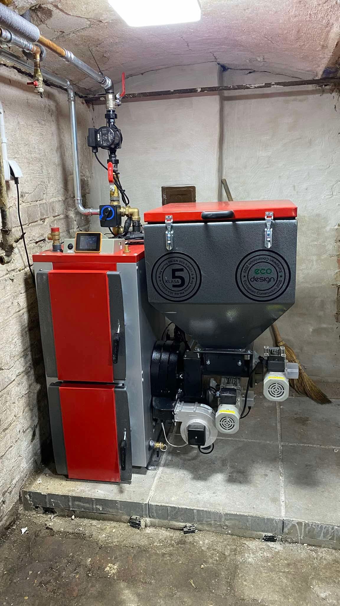 Czerwono-szary piec na paliwo stałe z podajnikiem, z widocznymi elementami instalacji grzewczej w kotłowni. Piec z certyfikatem Eco Design i klasą 5.