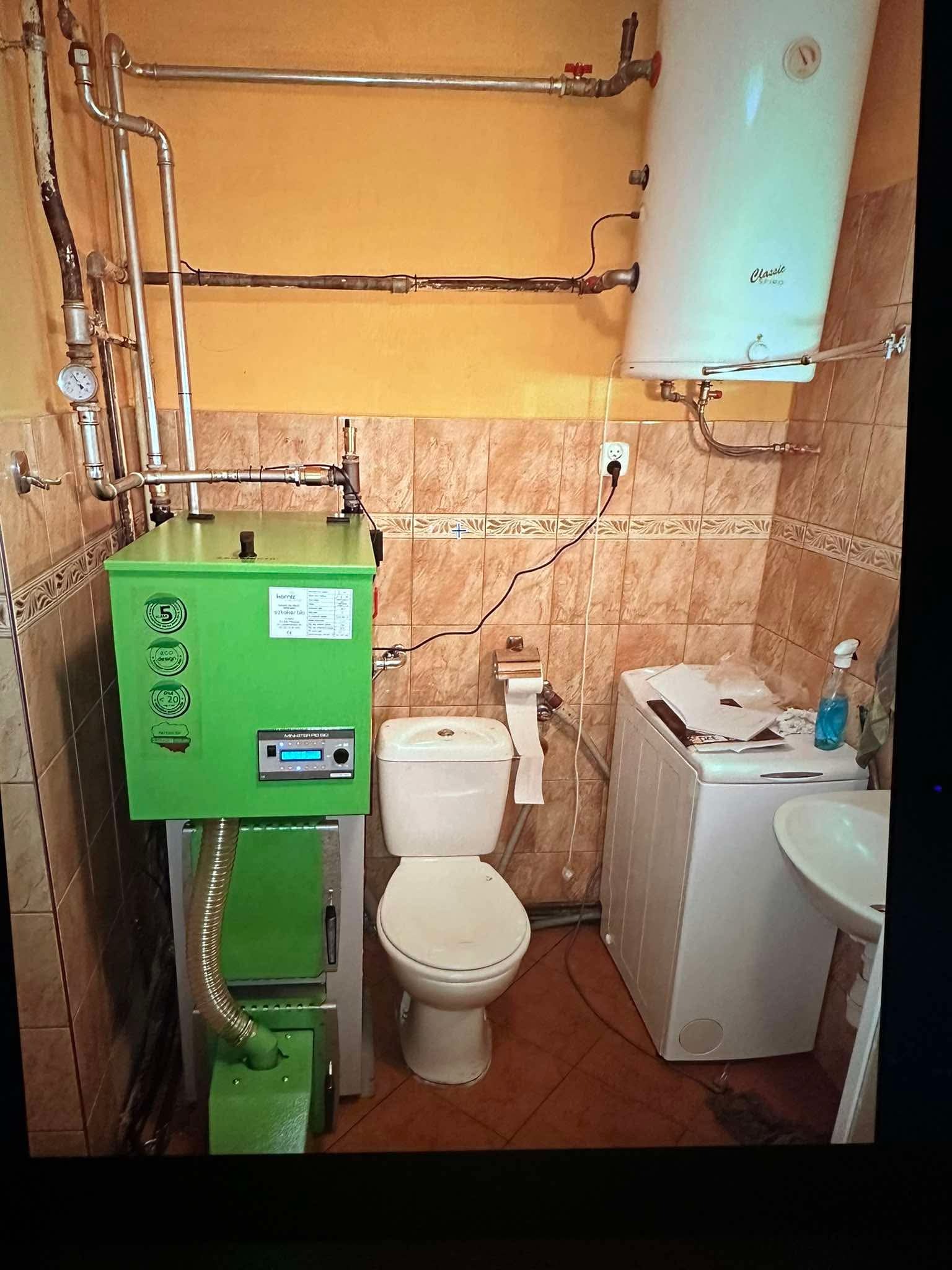 Zielony piec na paliwo stałe w małej łazience z brązowymi płytkami, obok toaleta i pralka. Widoczne rury instalacji grzewczej i bojler.