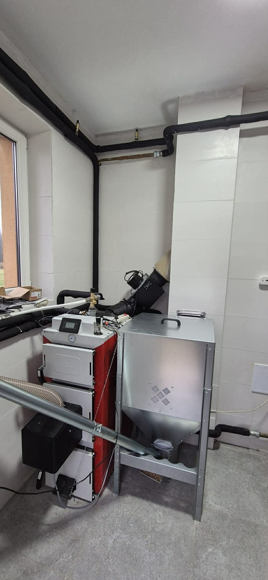 Nowoczesny piec na paliwo stałe z podajnikiem w kotłowni. Widoczne rury grzewcze z izolacją, białe płytki na ścianach i szary beton na podłodze.