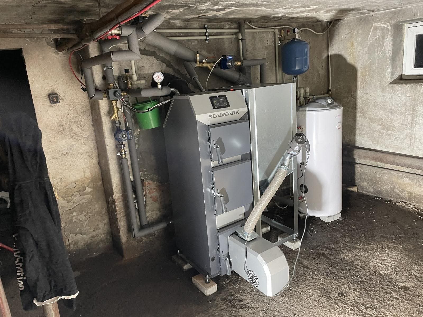 Szary piec na paliwo stałe marki Stalmark w piwnicy, z widocznymi rurami, zbiornikiem wyrównawczym i bojlerem. Instalacja grzewcza w surowym pomieszczeniu.