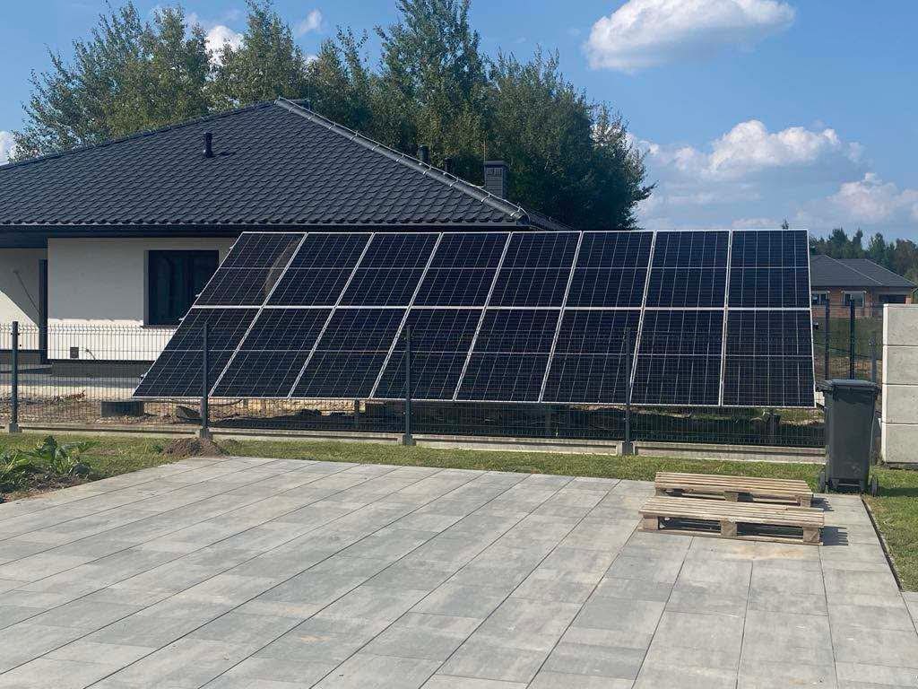 Panele fotowoltaiczne zamontowane na gruncie, widoczne z boku domu z ciemnym dachem. Na pierwszym planie szara kostka brukowa i palety. Słoneczny dzień.