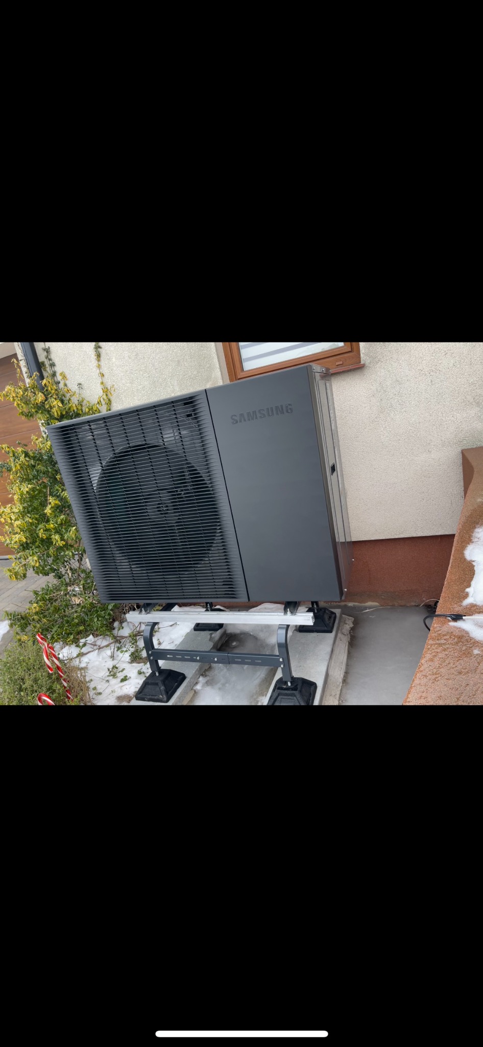 Zamontowana, czarna pompa ciepła Samsung na zewnątrz budynku, na specjalnej konstrukcji wsporczej. Widoczne elementy elewacji, roślinność i fragmenty dekoracji świątecznych.