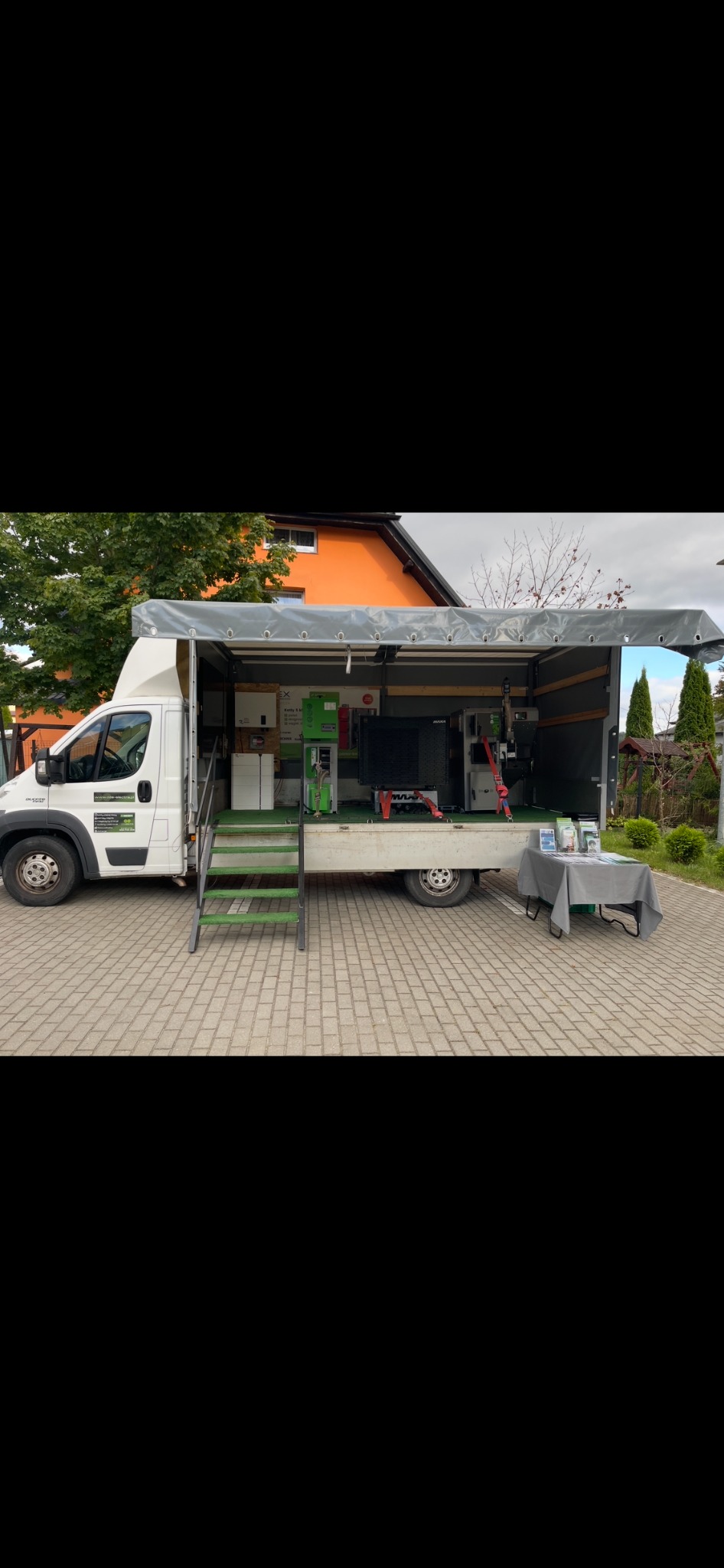 Mobilny punkt prezentacji: biały van z otwartą przestrzenią ładunkową, w której prezentowane są urządzenia grzewcze i informacyjne materiały na stole. Schodki z zieloną wykładziną.