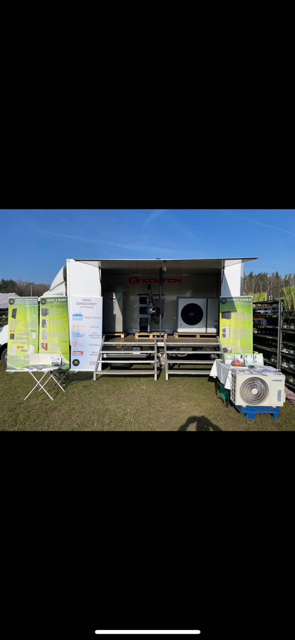 Mobilne stoisko z pompami ciepła i kotłami na targach. Prezentacja urządzeń marki Kołton, ulotki informacyjne, klimatyzator Samsung na palecie. Błękitne niebo w tle.