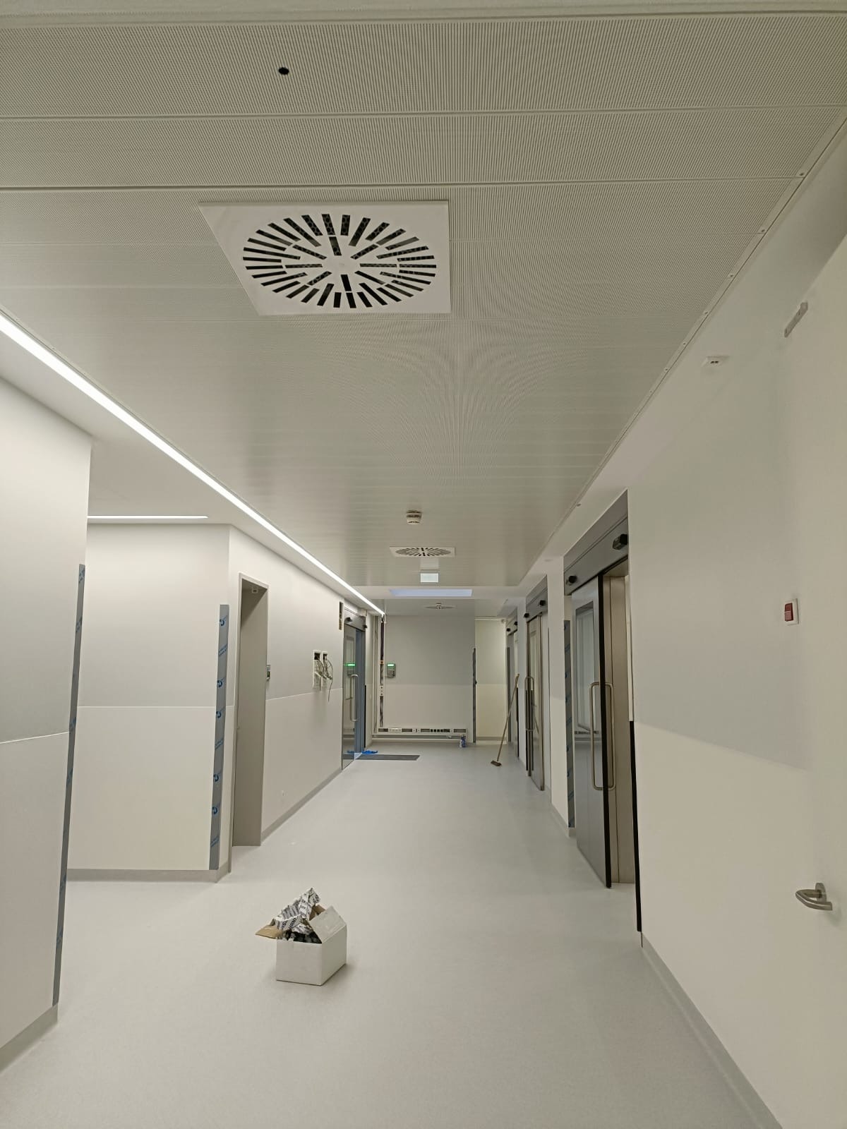 Długi, jasny korytarz z podwieszanym sufitem z kratką wentylacyjną i liniowym oświetleniem LED. Widoczne drzwi przesuwne i białe ściany. Na podłodze karton z materiałami.