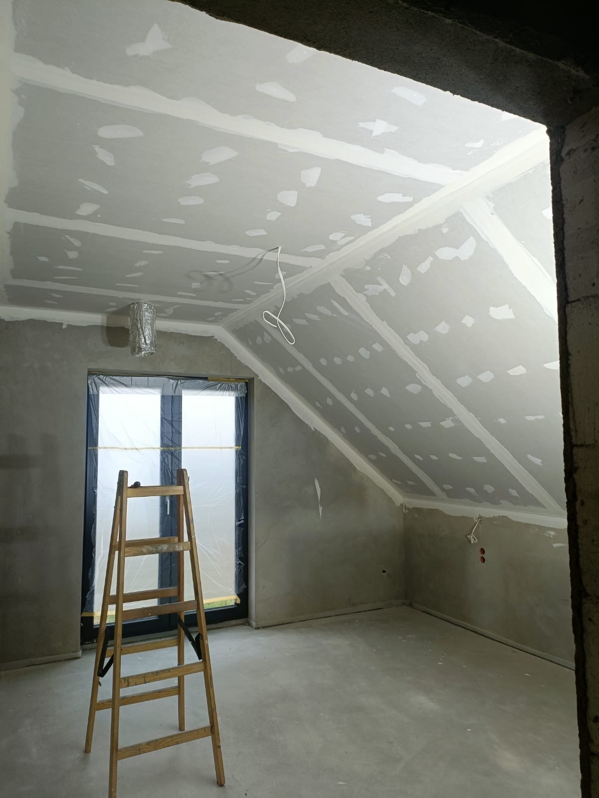 Skośny sufit po szpachlowaniu w Kokoszkowach, widoczne łączenia płyt gipsowych i drabina oparta o okno. Ściany surowe, przygotowane do dalszych prac.
