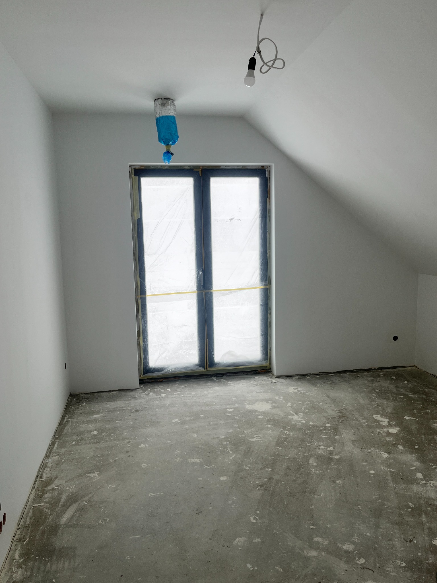 Pokój na poddaszu w trakcie prac wykończeniowych: świeżo pomalowane ściany na biało, okno zabezpieczone folią, surowa podłoga. Widoczne gniazdka elektryczne.
