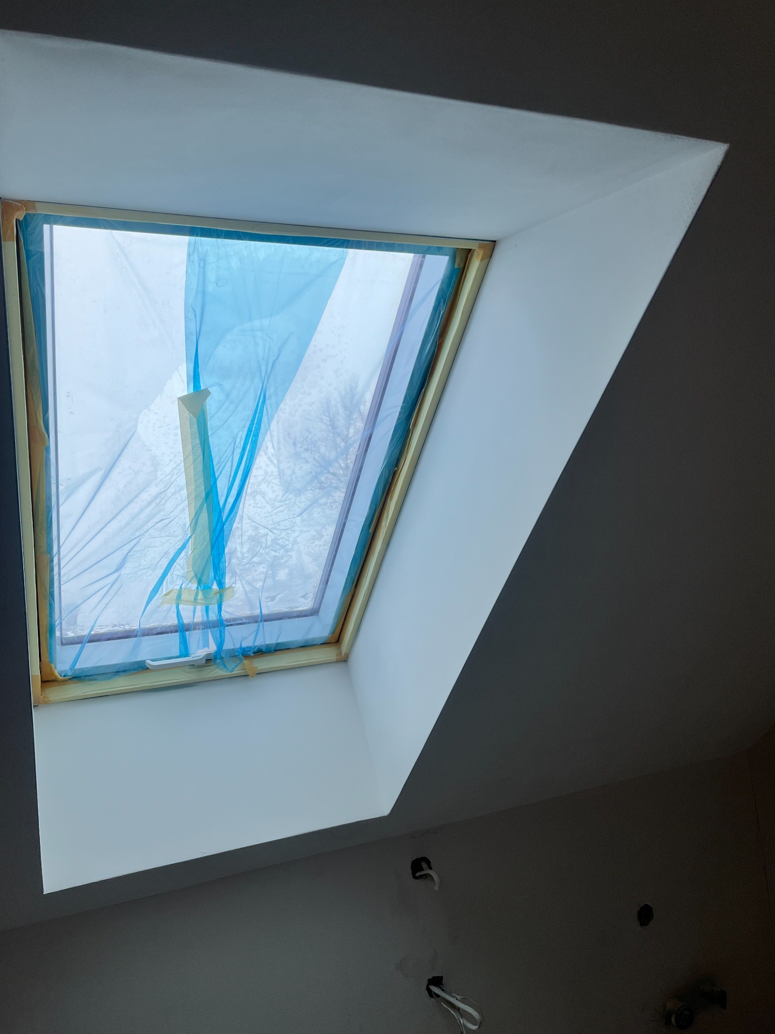 Okno dachowe zabezpieczone folią malarską, świeżo pomalowana wnęka okienna w odcieniach bieli, widoczne niedokończone instalacje elektryczne w szarej ścianie.