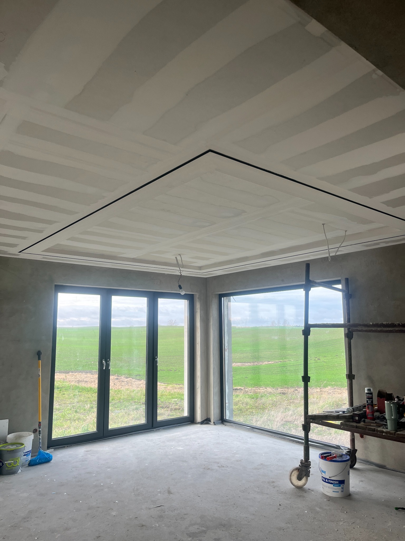 Pokój w trakcie szpachlowania sufitu z widocznymi łączeniami płyt gipsowych. Duże okna z widokiem na zielone pole. Rusztowanie i narzędzia malarskie w pomieszczeniu.