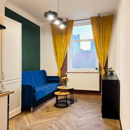 Kryspin Kozlowski Wykonczenia Wnetrz - Salon po remoncie: parkiet w jodełkę, niebieska sofa, żółte zasłony, ciemnozielona ściana. Widok na okno z ceglaną wnęką. Nowoczesne lampy wiszące.