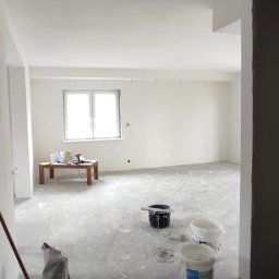 Kryspin Kozlowski Wykonczenia Wnetrz - Wnętrze w trakcie prac wykończeniowych: białe ściany i sufit, widoczne narzędzia malarskie, worki z zaprawą i wiaderka z farbą. Przy oknie stół z narzędziami.