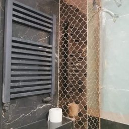 Kryspin Kozlowski Wykonczenia Wnetrz - Częściowo wykończona łazienka z grzejnikiem drabinkowym, czarnymi płytkami imitującymi marmur i dekoracyjną mozaiką w kształcie rybiej łuski na filarze.