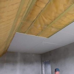 Kryspin Kozlowski Wykonczenia Wnetrz - Montaż płyty karton gips na poddaszu. Widoczne ocieplenie i konstrukcja nośna. Częściowo zamontowane płyty, surowe ściany. Laserowy poziom wyznacza linię.