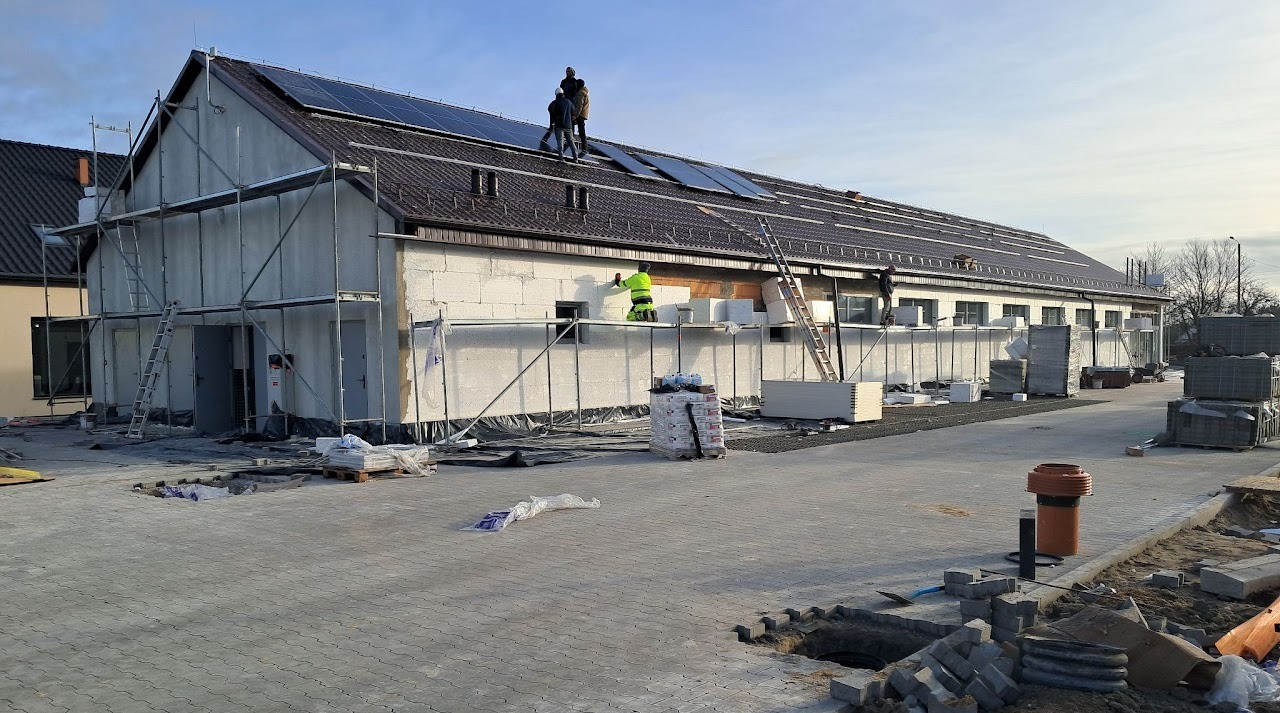 Budynek w trakcie budowy z panelami słonecznymi na dachu. Pracownicy na rusztowaniach i dachu. Ułożona kostka brukowa na placu. Widoczne materiały budowlane.