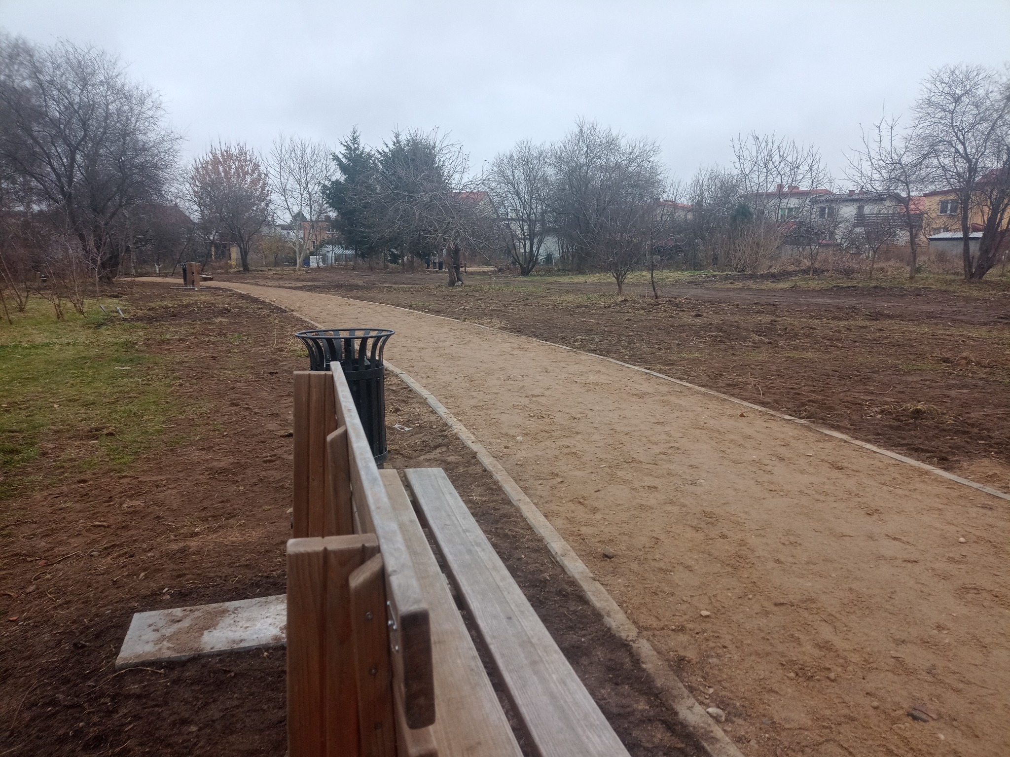 Ścieżka z piasku w parku, z ławką i koszem na śmieci po lewej stronie. W tle drzewa bez liści i budynki mieszkalne w pochmurny dzień.