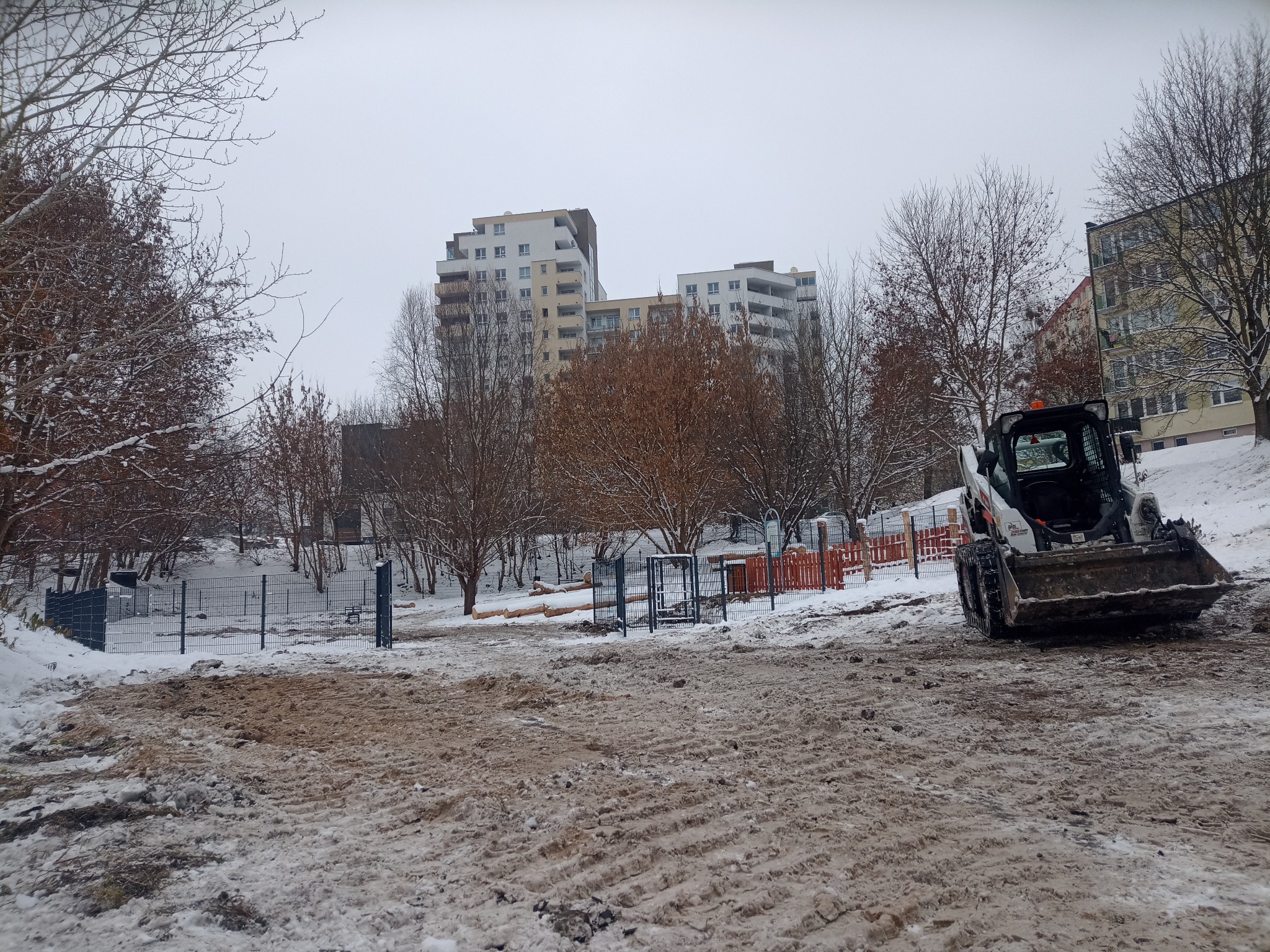 Zimowy krajobraz z miniładowarką Bobcat na placu budowy ogrodzenia panelowego. W tle bloki mieszkalne i drzewa bez liści, pokryte śniegiem. Szaro-biała kolorystyka.