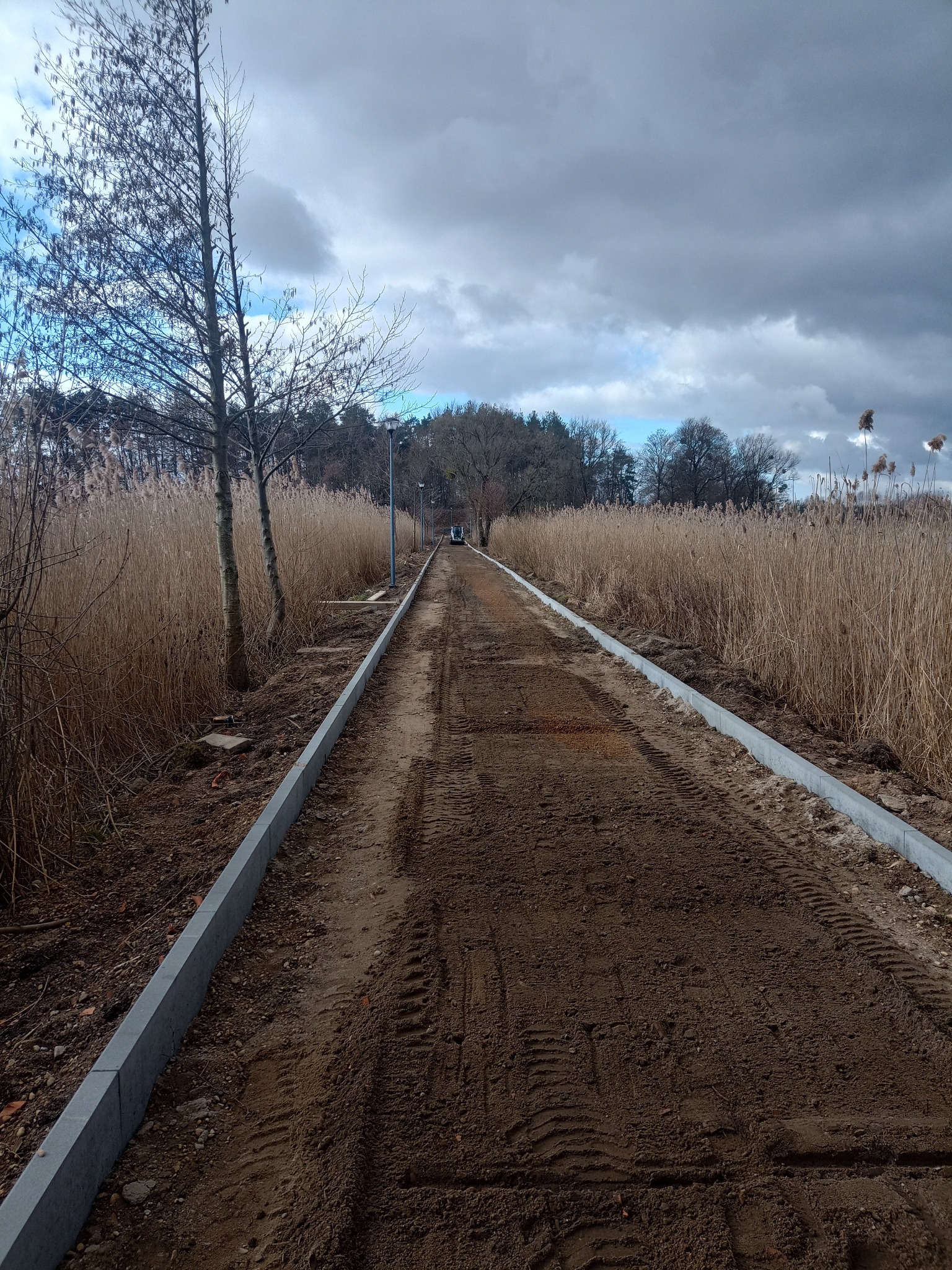 Przygotowany teren pod ścieżkę z obrzeżami, widoczne ślady opon, tło z trzcinami i drzewami, pochmurne niebo. Prace ziemne w toku.