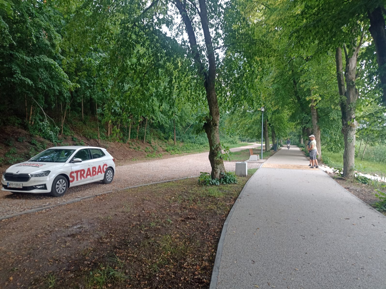 Biały samochód firmy Strabag zaparkowany obok ścieżki rowerowej w parku. W tle widoczni ludzie i drzewa. Ścieżka wykonana z szarej nawierzchni.