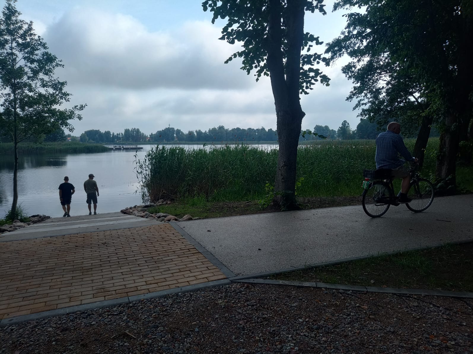Brzeg jeziora z zejściem z kostki brukowej, ścieżka rowerowa i spacerujący ludzie. Człowiek na rowerze jedzie ścieżką wzdłuż brzegu.