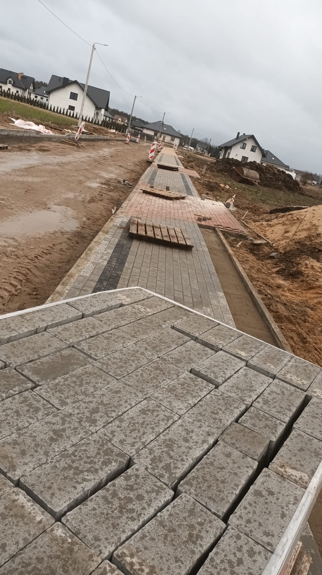 Ułożona część chodnika z kostki brukowej w nowym osiedlu. Na pierwszym planie paleta z kostką. Widoczne domy w tle i przygotowany teren pod dalsze prace.