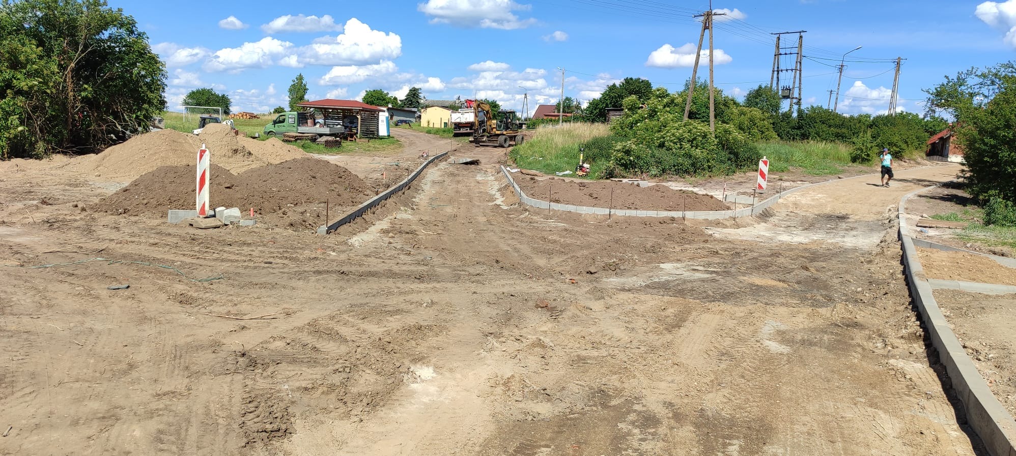 Etap budowy drogi gruntowej z obrzeżami betonowymi, widoczny sprzęt budowlany i hałdy ziemi. Słoneczny dzień, niebieskie niebo z chmurami.