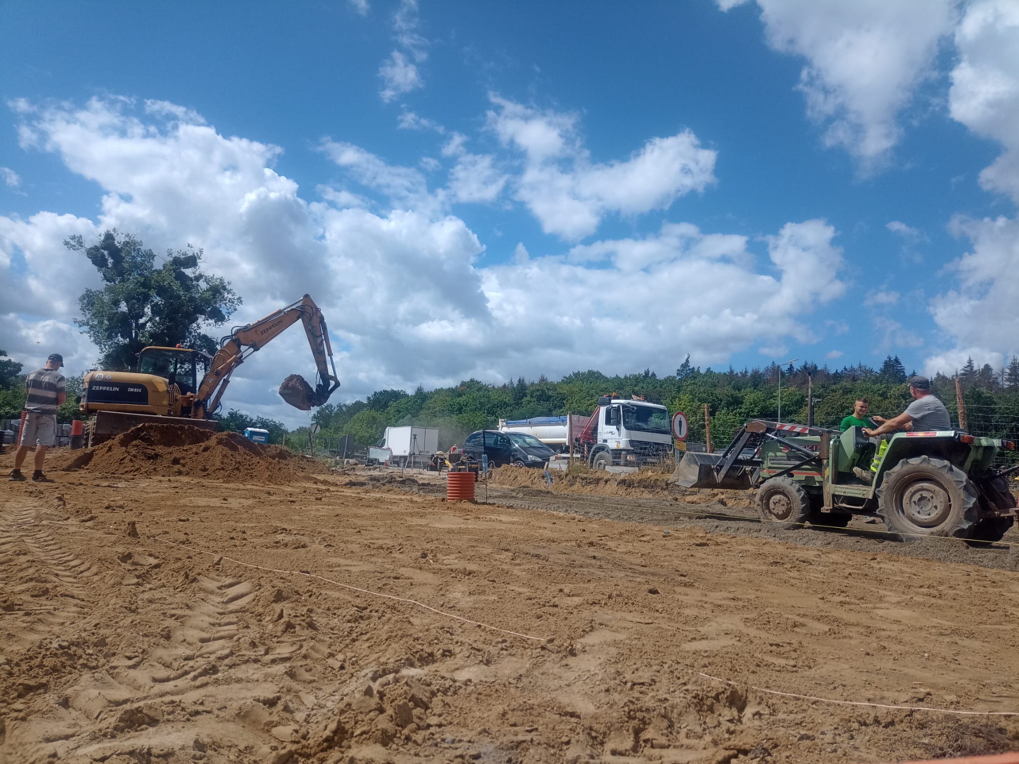 Żółta koparka Zeppelin i traktor z łyżką na placu budowy, przygotowany teren pod budowę drogi, widoczne ciężarówki i pracownicy, błękitne niebo z chmurami.