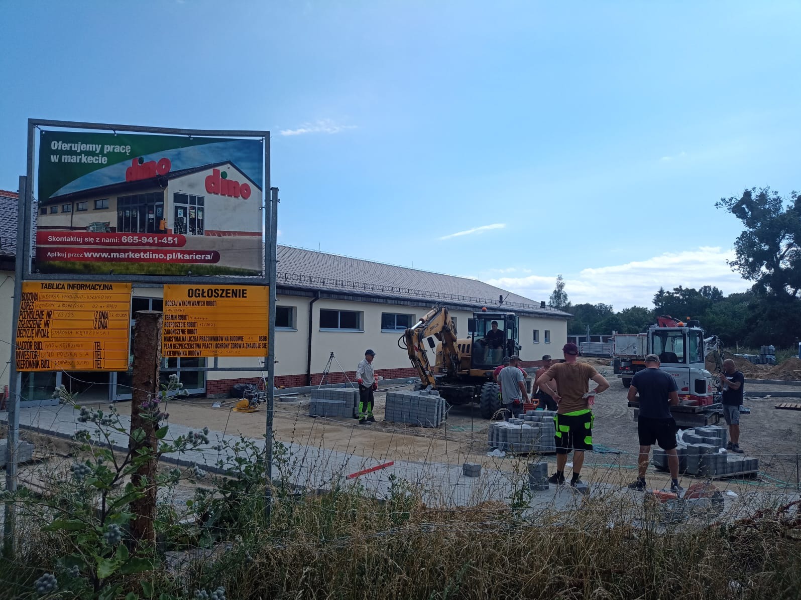 Budowa parkingu przed marketem Dino: ekipa budowlana, maszyny (koparka, wywrotka), palety z kostką brukową. Tablica informacyjna budowy i reklama Dino w tle.