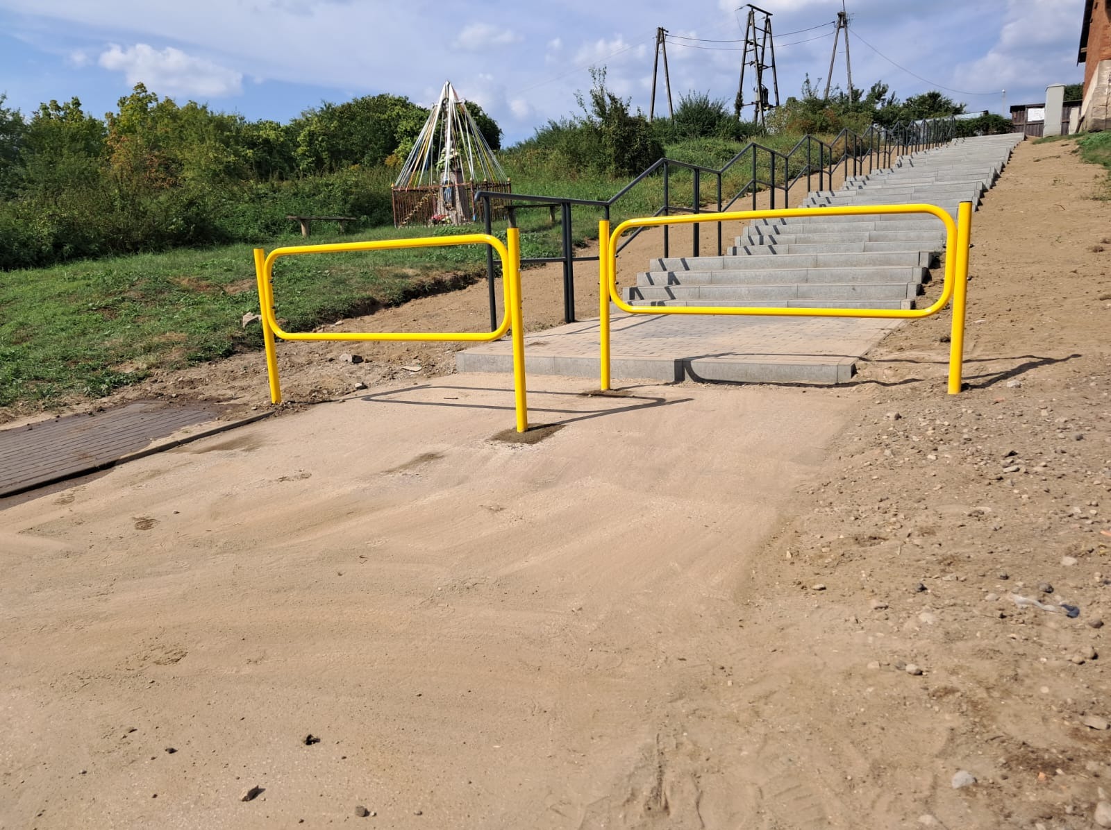 Nowe schody z barierkami w przestrzeni publicznej. Żółte barierki na wejściu, czarne poręcze schodów. Widok na górę, w tle zieleń i elementy infrastruktury.