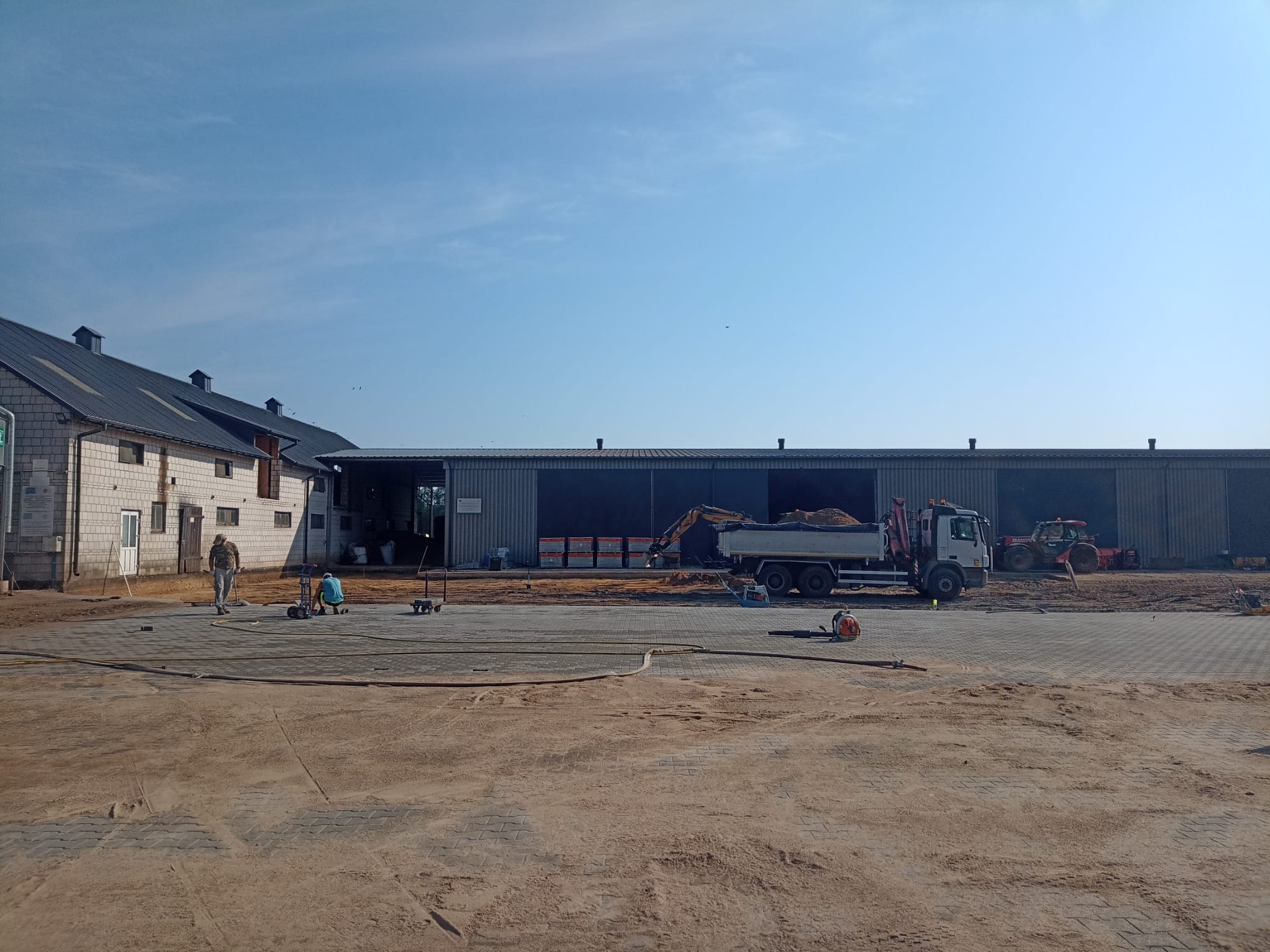 Prace brukarskie na dużej powierzchni: robotnicy, maszyny (koparka, ciężarówka, zagęszczarka), częściowo ułożona kostka. Budynek gospodarczy w tle, błękitne niebo.