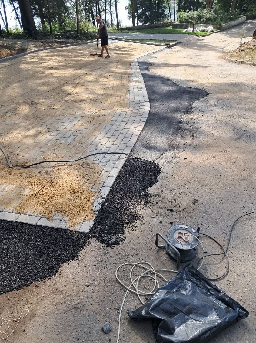 Układanie kostki brukowej na ścieżce w parku. Widoczny fragment ułożonej kostki, piasek, oraz osoba zamiatająca powierzchnię. W tle drzewa i zieleń.