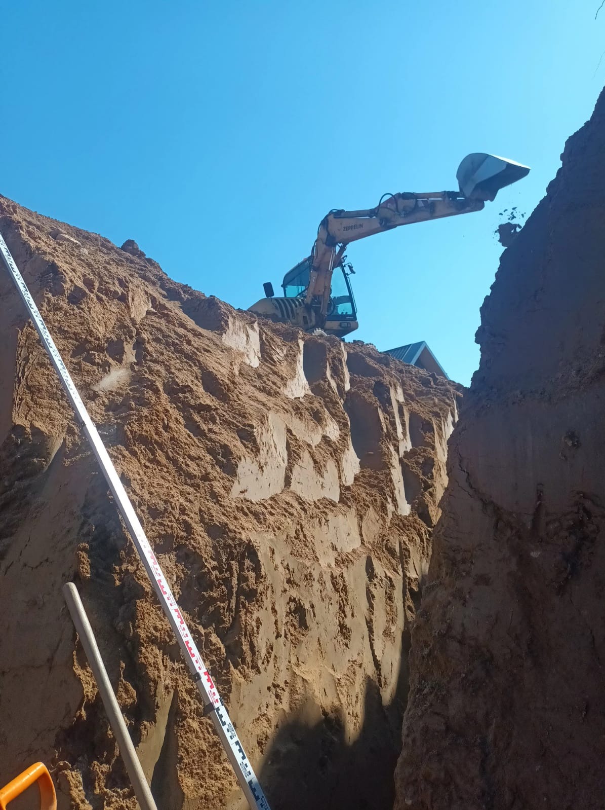 Żółta koparka Zeppelin w trakcie prac ziemnych, widoczna z dołu wykopu. W tle błękitne niebo, a w kadrze miarka geodezyjna i fragmenty narzędzi.