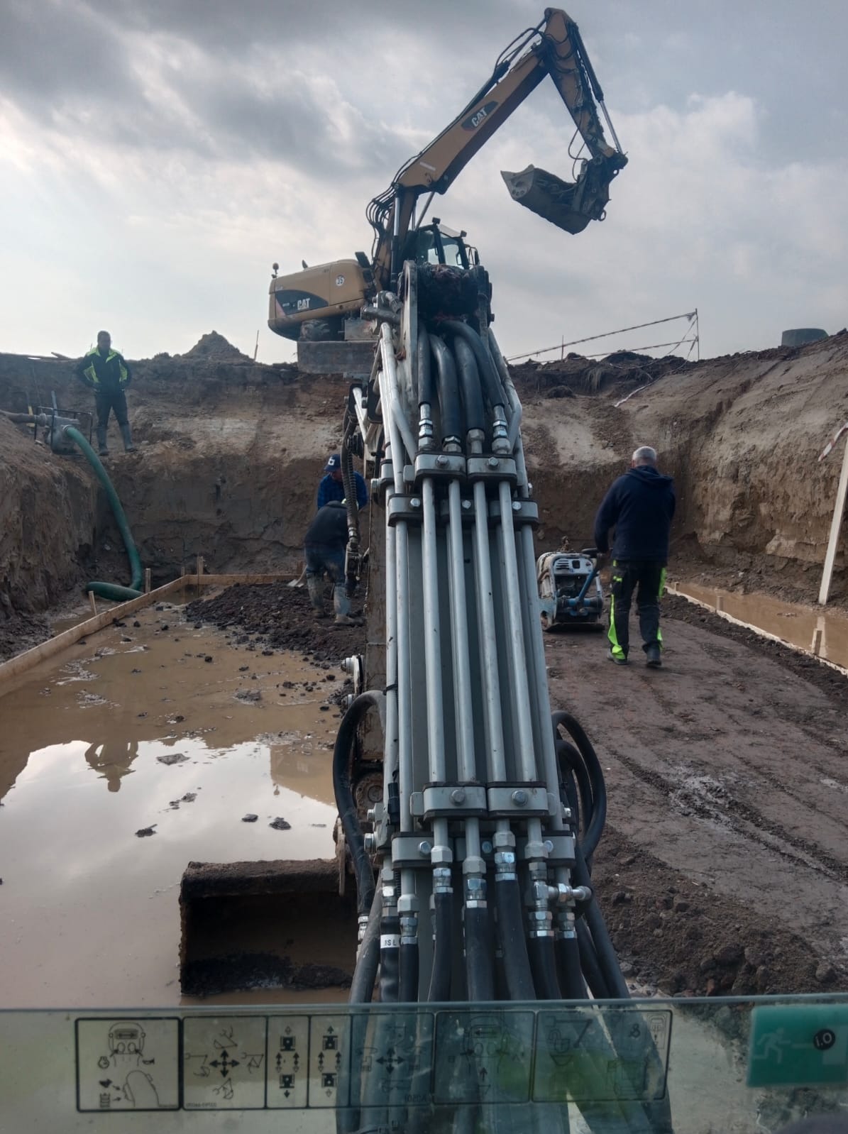 Żółta koparka CAT w trakcie prac ziemnych. Widok z kabiny operatora na wykop, pracowników i sprzęt. W tle pochmurne niebo.