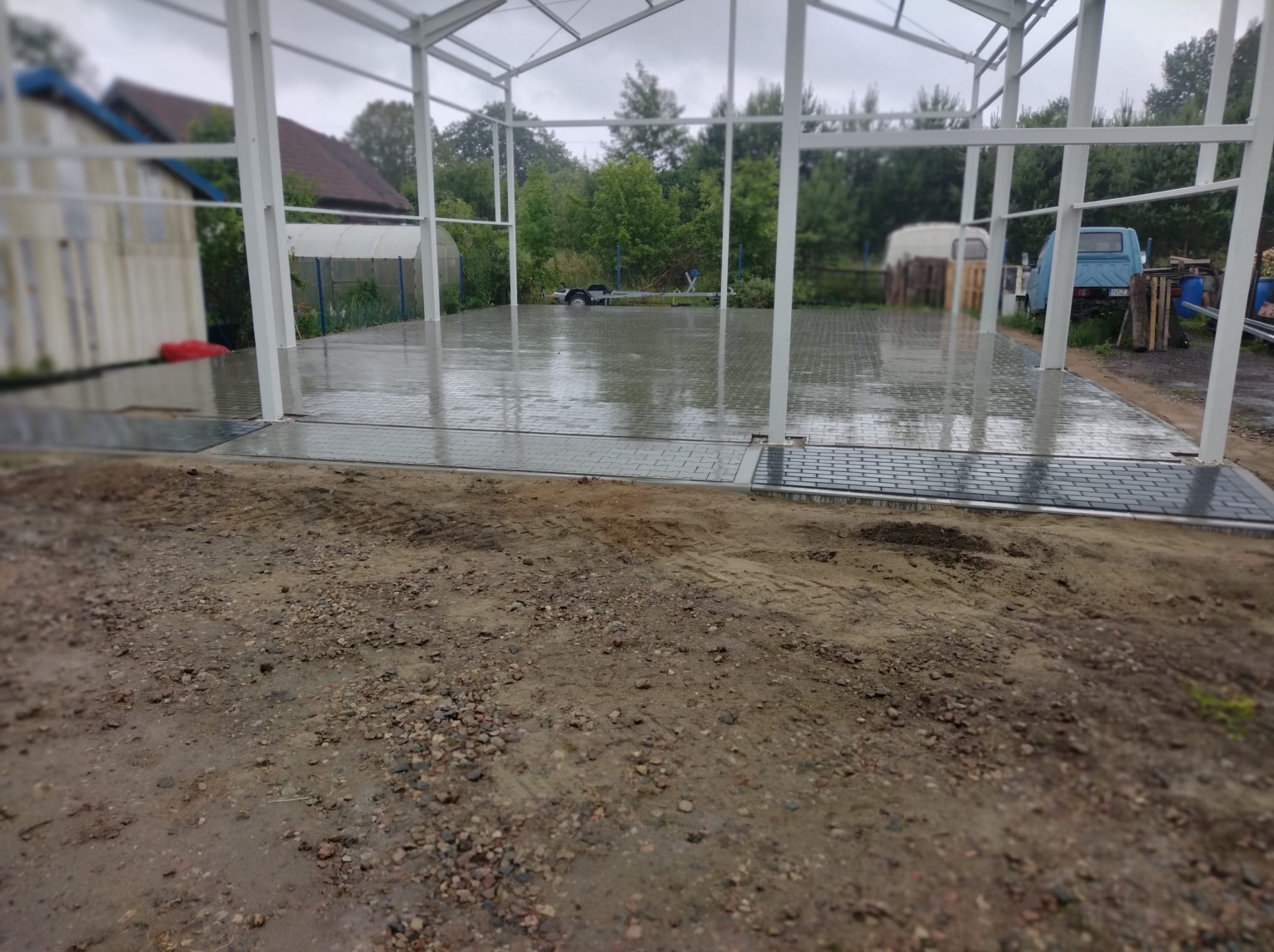 Szara kostka brukowa pod zadaszeniem z metalową konstrukcją. Na kostce widoczne odbicia wody. W tle zieleń drzew i zabudowania. Ziemia przed kostką częściowo mokra.
