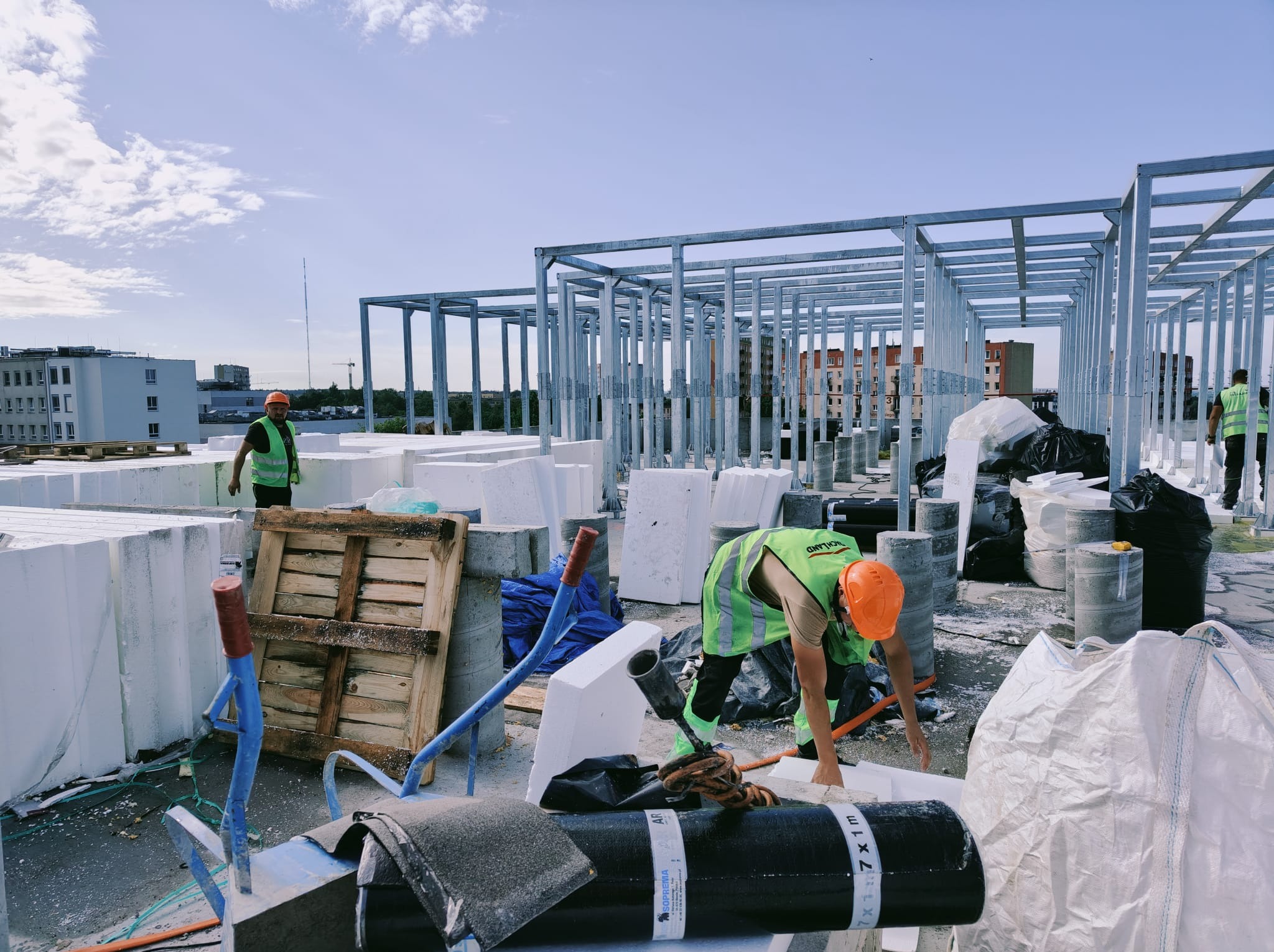 Prace budowlane na dachu: robotnicy w kaskach i kamizelkach, stalowa konstrukcja, białe płyty izolacyjne, materiały budowlane. Widok na budynki w tle.