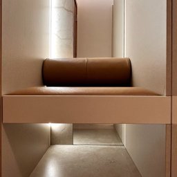 SPACE ARCHITECTS - Wnęka z siedziskiem z brązowej skóry, oświetlona LED, w zabudowie z beżowych paneli. Widok na korytarz z lustrzanym odbiciem pod siedziskiem. Minimalistyczny design.