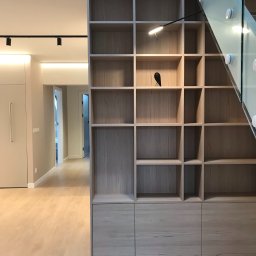 SPACE ARCHITECTS - Jasna, drewniana biblioteczka z oświetleniem punktowym i dekoracyjną figurką ptaka, zintegrowana ze szklaną balustradą schodów w nowoczesnym wnętrzu.