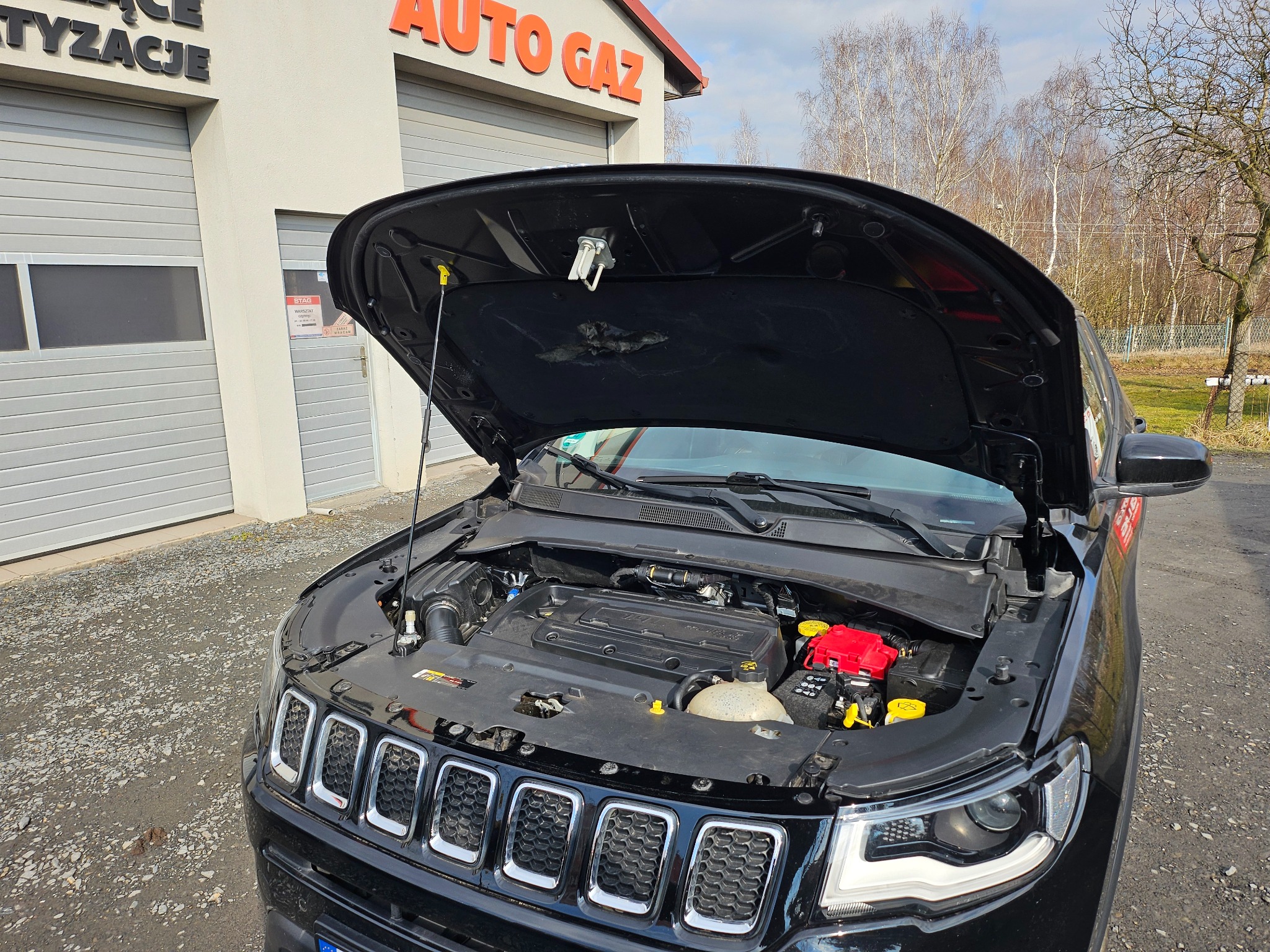 Czarny Jeep Compass z otwartą maską na tle budynku warsztatu Auto Gaz. Widoczny silnik i elementy instalacji. Ujęcie z bliska, z zewnątrz.