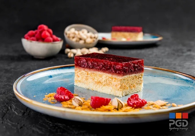 Kremowy torcik z warstwą malinowej galaretki na błękitnym talerzu, udekorowany malinami i pistacjami. W tle miseczki z malinami i orzechami.