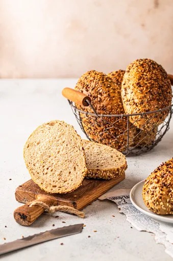 Kosz pełen bułek z ziarnami, kromki na desce i nóż z ząbkami. Jasne tło, naturalne światło. Bułki rustykalne, idealne do kanapek.