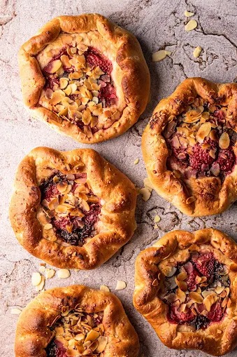 Rustykalne galette z owocami leśnymi i płatkami migdałów na szorstkim, kamiennym tle. Cztery okrągłe ciastka z widocznym nadzieniem.