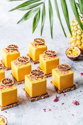 Żółte kostki deserowe na krakersach, udekorowane suszoną pomarańczą i płatkami róż, z ananasem i marakują w tle, pod liściem palmy. Eleganckie i apetyczne.
