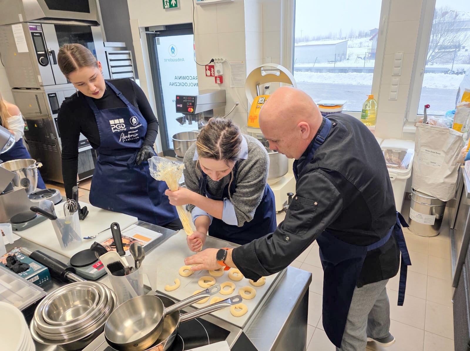 Zespół w kuchni formuje ciasto na pączki z worka cukierniczego na blasze. W tle okno z zimowym krajobrazem i profesjonalny sprzęt kuchenny.