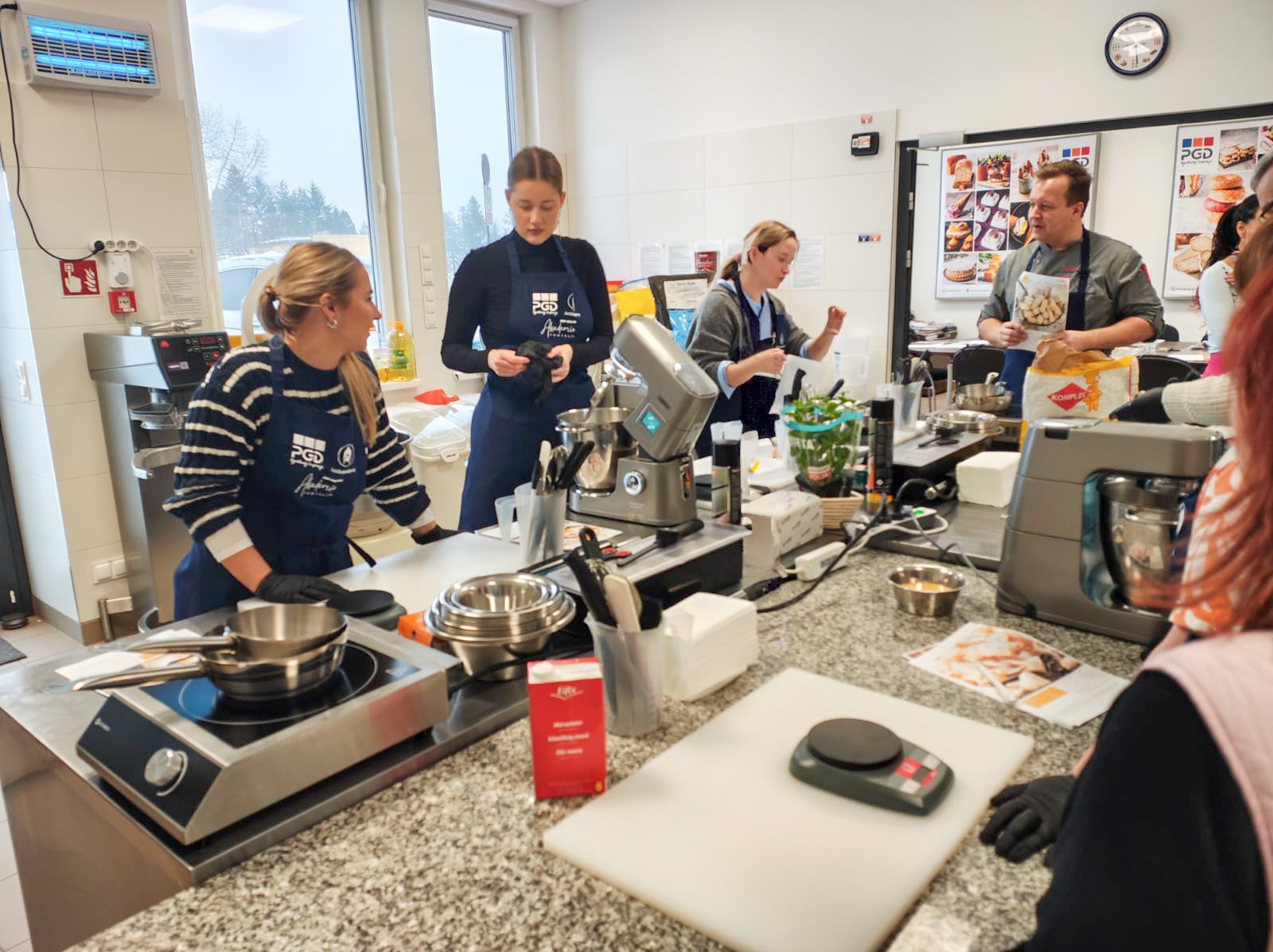 Warsztaty kulinarne: grupa osób w fartuchach przygotowuje potrawy w profesjonalnej kuchni z nowoczesnym sprzętem, w tle plakaty z daniami.