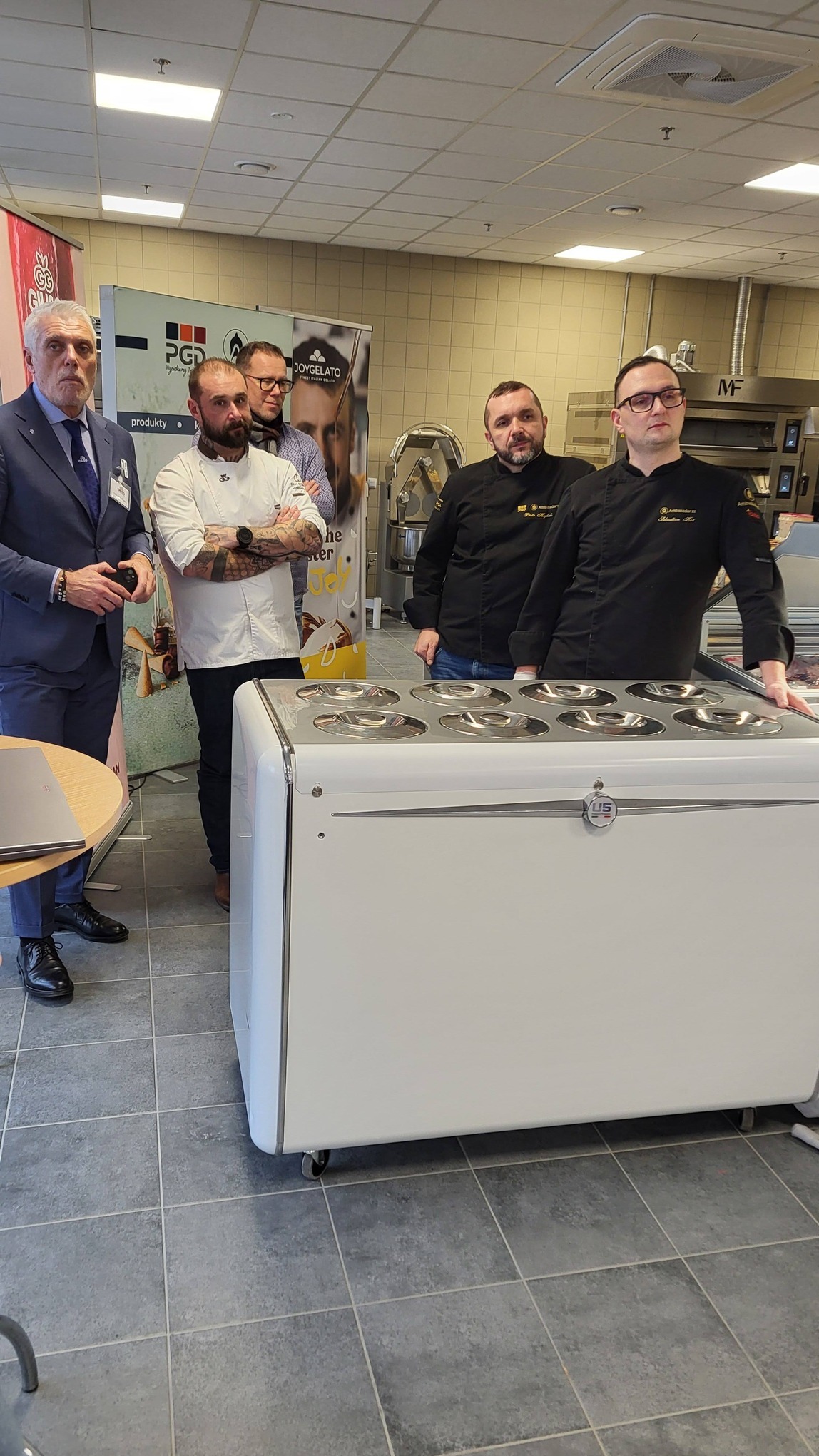 Prezentacja profesjonalnego sprzętu kuchennego z kucharzami w tle, w jasnym pomieszczeniu z szarymi płytkami podłogowymi i banerami reklamowymi.