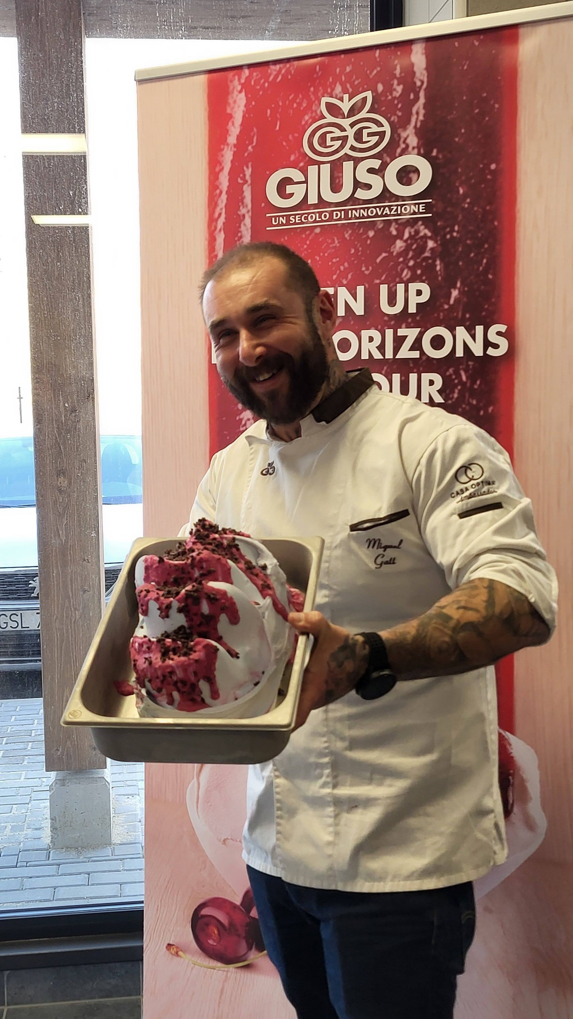 Uśmiechnięty kucharz w białym uniformie prezentuje metalową formę z lodami o smaku owoców leśnych, na tle baneru reklamowego marki lodów.