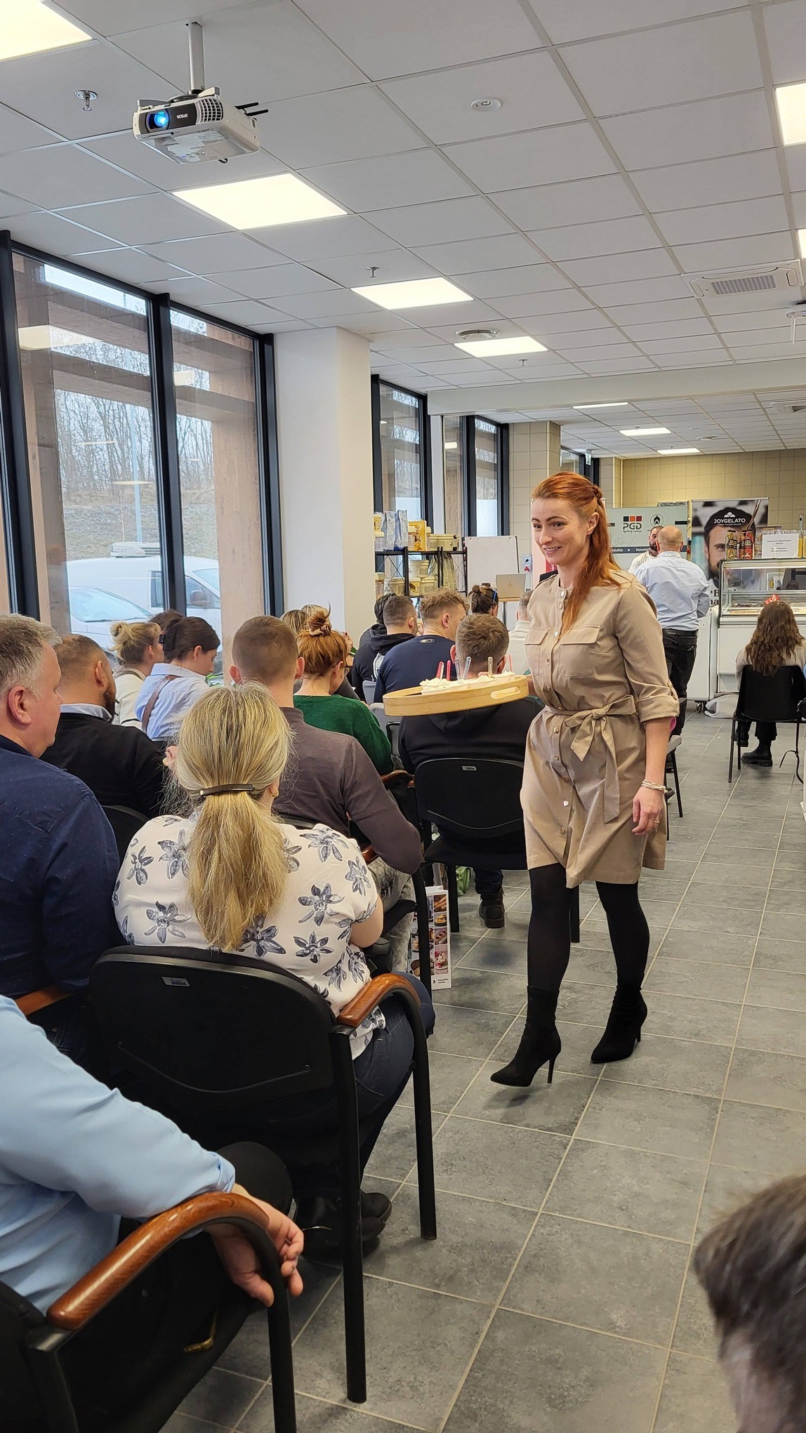 Kobieta w sukience niesie tacę z przekąskami w sali konferencyjnej, gdzie siedzą ludzie. W tle widoczna kuchnia i okna.