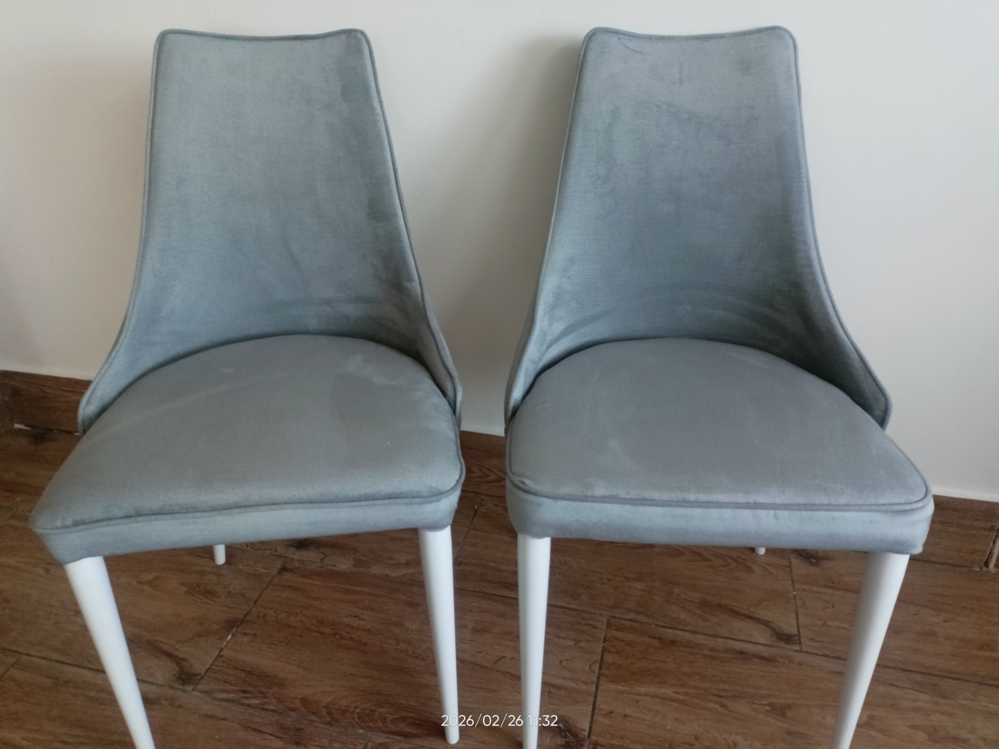 Dwa krzesła tapicerowane szarą tkaniną, z białymi nogami, ustawione obok siebie na tle jasnej ściany. Widoczne detale wykończenia i faktura materiału.